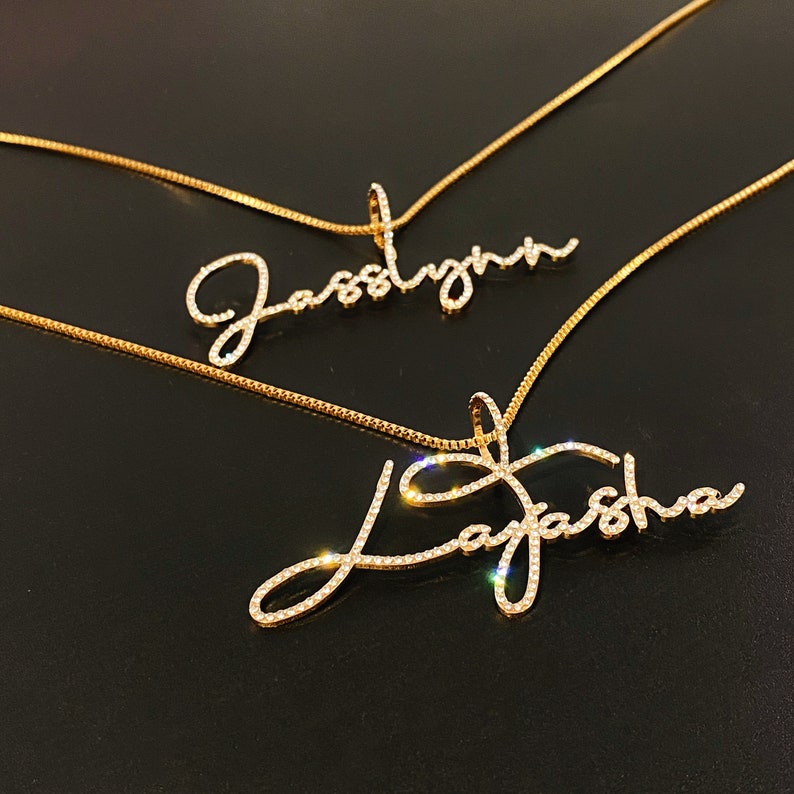 May include: Two gold-colored name necklaces. One necklace spells out "Jasslynn" and the other spells out "Layasha". The names are written in a cursive font and are encrusted with small, sparkling crystals. The necklaces are on a dark background.