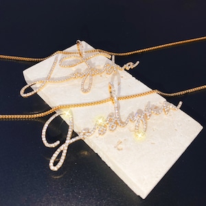 May include: Two gold-colored name necklaces, each spelling out a name in cursive script, encrusted with small, sparkling crystals. The necklaces rest on a white, rectangular surface against a dark background. The chains are also gold.