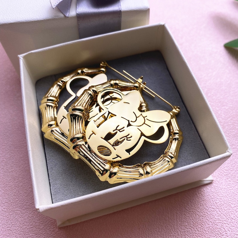 May include: Gold hoop earrings with a cutout design of a cartoon character's face. The earrings are in a white box with a grey interior.