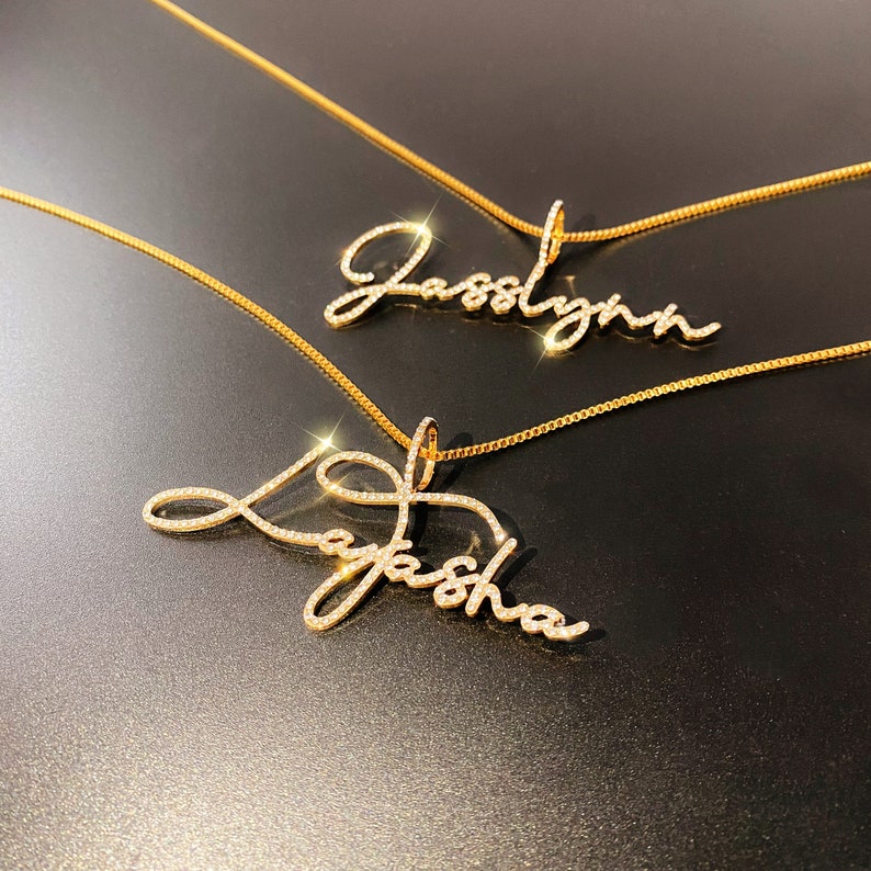 May include: Two gold-colored name necklaces with diamond-like embellishments. One necklace spells out "Jasslynn" and the other says "Layasha" in a cursive font. The necklaces are displayed on a dark, reflective surface.