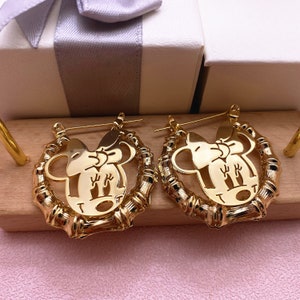 May include: Gold hoop earrings with a cutout design of a cartoon mouse with a bow. The earrings are made of a bamboo-like material.