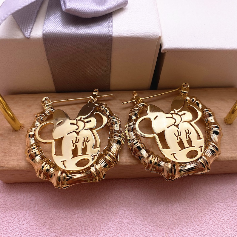 May include: Gold hoop earrings with a cutout design of a cartoon mouse with a bow. The earrings are made of a bamboo-like material.