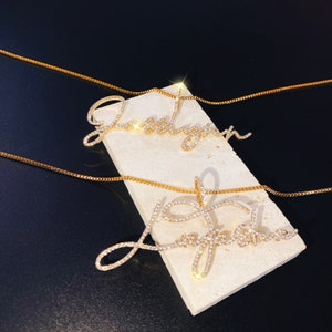 May include: Two gold-colored name necklaces, each spelling a different name in cursive script. The names are encrusted with small, clear crystals. The necklaces rest on a rectangular, off-white surface against a dark background.