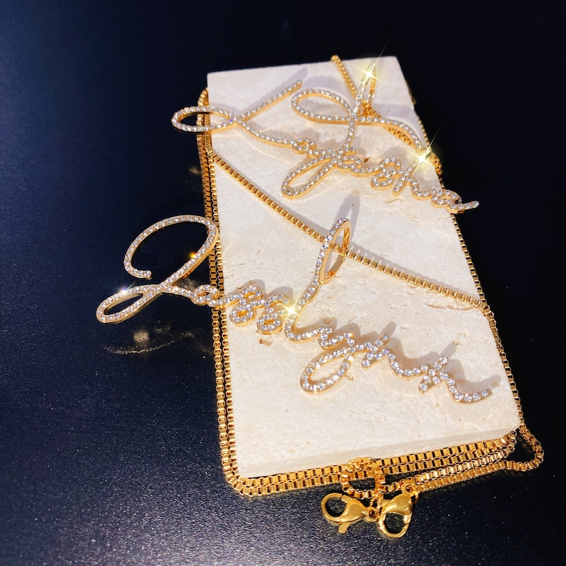 May include: Gold-toned necklaces with the word "Joy" spelled out in cursive script, embellished with small, sparkling crystals. The necklaces are displayed on a rectangular, off-white surface against a dark background. The chains have a delicate, box-link design.