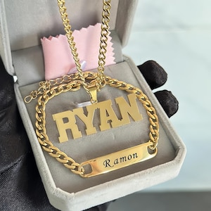 May include: Gold-coloured necklace and bracelet set. The necklace pendant spells out "RYAN" in block letters. The bracelet has the name "Ramon" engraved. The jewellery is displayed in a grey box with a pink cloth.