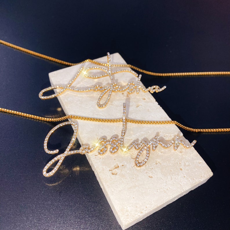 May include: Two gold-colored name necklaces, each spelling "Jesslyn" in a cursive font, are displayed on a light-colored rectangular block. The letters are encrusted with small, sparkling crystals. The necklaces rest on a dark background.