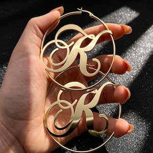 May include: A pair of large gold-toned hoop earrings featuring a polished finish. Each circular hoop contains a prominent, stylized cursive letter K centered within the frame. The design is crafted from a smooth, reflective metal material.