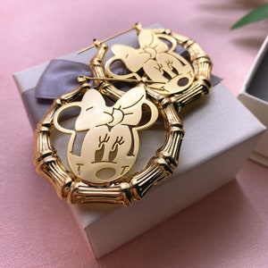 May include: Gold hoop earrings with a cutout design of a cartoon character with a bow. The earrings are made of bamboo-like material.