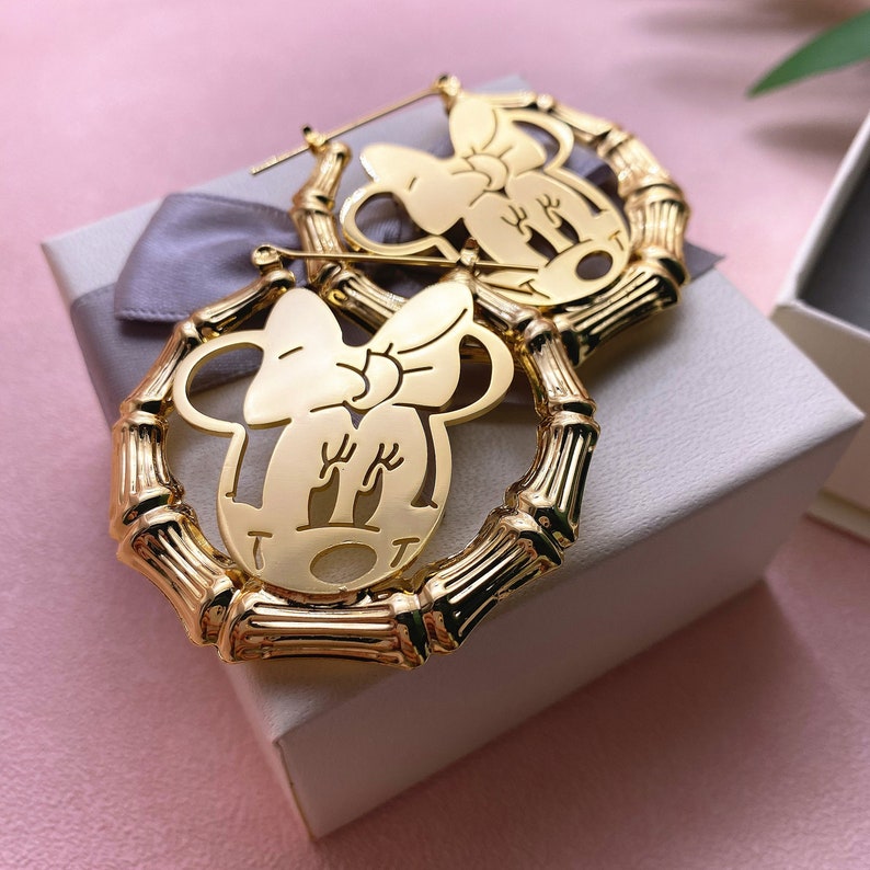 May include: Gold hoop earrings with a cutout design of a cartoon character with a bow. The earrings are made of bamboo-like material.