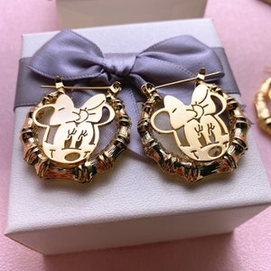 May include: Gold-tone earrings with a bamboo-style design and a cutout of a cartoon character with a bow. The earrings are displayed in a white gift box with a grey ribbon.