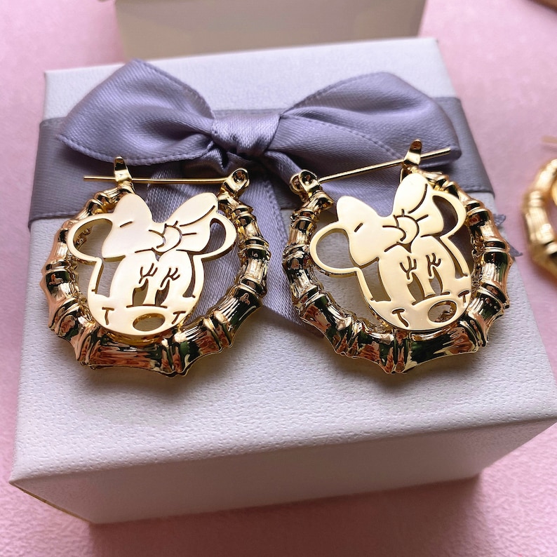 May include: Gold-tone earrings with a bamboo-style design and a cutout of a cartoon character with a bow. The earrings are displayed in a white gift box with a grey ribbon.