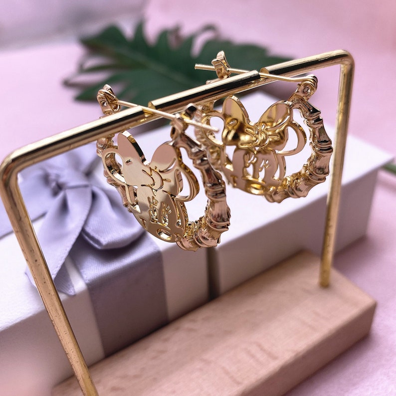 May include: A pair of gold hoop earrings with a bow design. The earrings are on a gold stand.