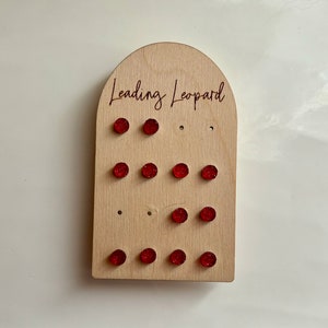 May include: A wooden earring display stand with the text "Leading Leopard". The stand holds several pairs of red glitter stud earrings. The earrings are arranged in rows on the light-coloured wood. The stand is arched at the top.