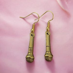 May include: Gold-tone microphone earrings with a hook closure. The microphone has a textured surface and a small 'B' engraved on the side.