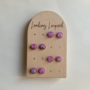 Peut inclure: Un présentoir en bois avec le texte "Leading Leopard" gravé dessus. Le présentoir comporte plusieurs trous, chacun contenant une paire de boucles d'oreilles en résine violette avec des paillettes dorées.