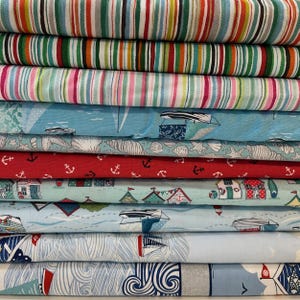 May include: A selection of patterned fabrics, stacked neatly. The top layers feature striped designs in colours such as red, green, and white. Beneath are fabrics with nautical motifs, including anchors, sailboats, and beach huts. The colour scheme includes red, blue, and white.