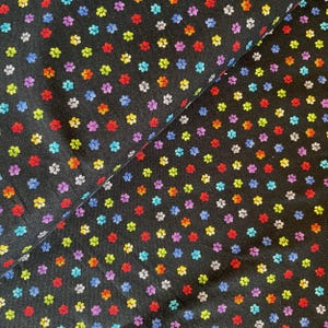 May include: Black fabric featuring a repeating pattern of colourful paw prints. The paw prints are in various colours, including red, orange, yellow, green, blue, and purple. The fabric is folded, showing the texture and pattern.