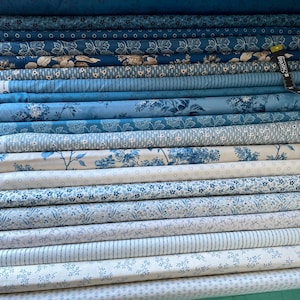 May include: A collection of stacked fabric bolts featuring various shades of blue and white. The patterns include floral motifs, geometric shapes, stripes, and polka dots. A black measuring tape is visible on the right side of the stack.