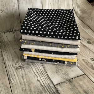 May include: A stack of folded fabric squares featuring various patterns including black with white polka dots, solid white, grey with yellow stars, black with white stars, yellow with white polka dots, and grey with white star motifs.