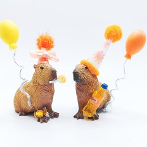 May include: Two capybaras wearing party hats. One capybara is holding a small orange and yellow lollipop. The other capybara is holding a small orange soda bottle. Both capybaras are wearing party hats. One hat is pink with white flowers and the other is orange with white polka dots.