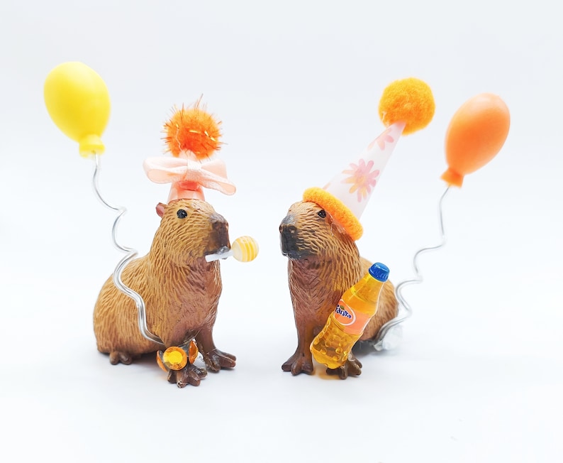 May include: Two capybaras wearing party hats. One capybara is holding a small orange and yellow lollipop. The other capybara is holding a small orange soda bottle. Both capybaras are wearing party hats. One hat is pink with white flowers and the other is orange with white polka dots.