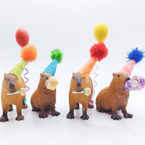 May include: Four capybara figurines wearing colourful party hats with a balloon, lollipop, and a swirl of paper on top. The capybaras are brown and have black eyes and noses.