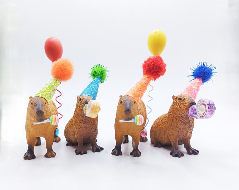 May include: Four capybara figurines wearing colourful party hats with a balloon, lollipop, and a swirl of paper on top. The capybaras are brown and have black eyes and noses.