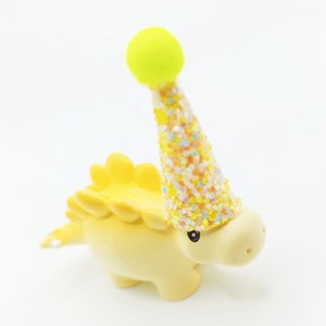 May include: A yellow dinosaur figurine wearing a yellow, pink, and blue confetti party hat with a yellow pom-pom on top.