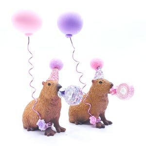 May include: Two brown capybara figurines wearing pink and purple party hats with pink and white confetti blowers. The capybaras are standing on a white surface with pink and purple balloons.