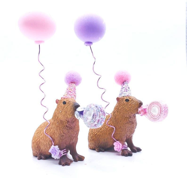 May include: Two brown capybara figurines wearing pink and purple party hats with pink and white confetti blowers. The capybaras are standing on a white surface with pink and purple balloons.