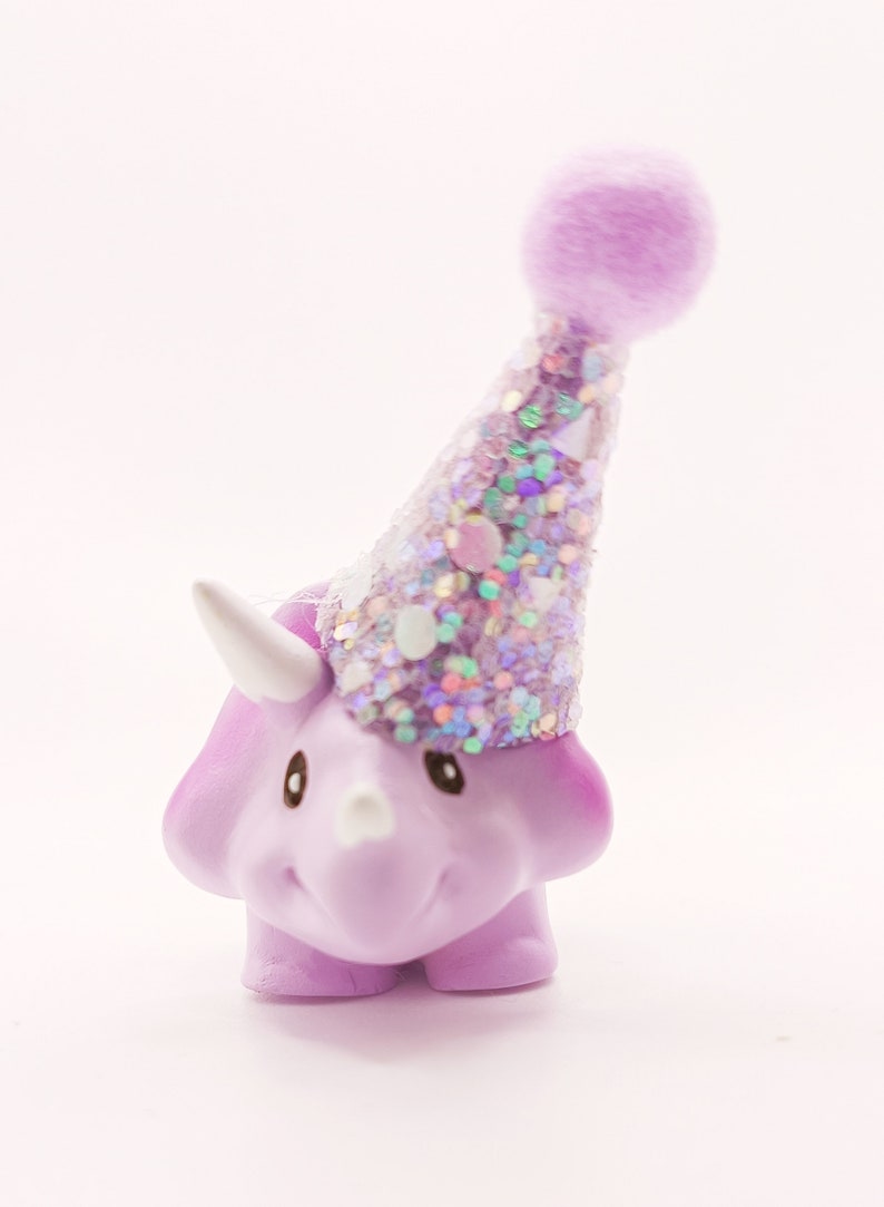 May include: A purple dinosaur figurine wearing a sparkly purple party hat with a fuzzy pom-pom on top.