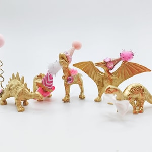 Puede incluir: Cuatro figuritas de dinosaurio pintadas de oro que llevan sombreros de fiesta rosas y blancos. Los dinosaurios son un estegosaurio, un triceratops, un pterod&aacute;ctilo y un braquiosaurio. Un dinosaurio tiene un globo rosa atado a su cola.