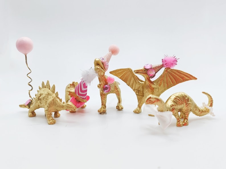 Puede incluir: Cuatro figuritas de dinosaurio pintadas de oro que llevan sombreros de fiesta rosas y blancos. Los dinosaurios son un estegosaurio, un triceratops, un pterod&aacute;ctilo y un braquiosaurio. Un dinosaurio tiene un globo rosa atado a su cola.