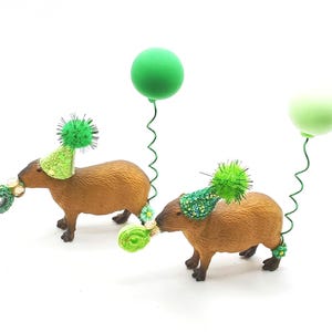 May include: Two brown capybara figurines wearing green party hats and holding green balloons. The capybaras are standing on a white surface.