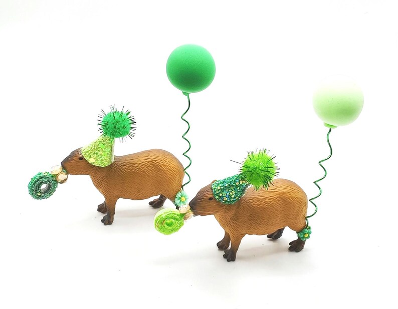 May include: Two brown capybara figurines wearing green party hats and holding green balloons. The capybaras are standing on a white surface.