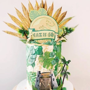 Puede incluir: Un pastel de cumplea&ntilde;os verde y dorado decorado con una silueta de dinosaurio, hojas de palma y una pancarta que dice "Caz tiene 60 a&ntilde;os". El pastel est&aacute; coronado con un logotipo de Jurassic Park y una peque&ntilde;a figura de dinosaurio.