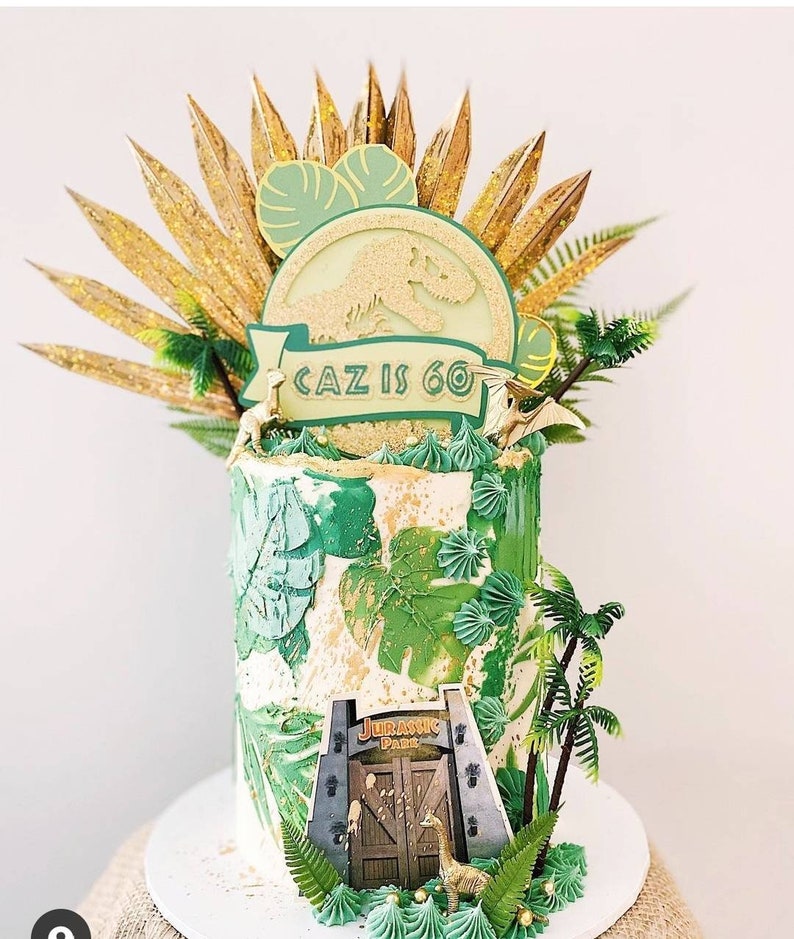 Puede incluir: Un pastel de cumplea&ntilde;os verde y dorado decorado con una silueta de dinosaurio, hojas de palma y una pancarta que dice "Caz tiene 60 a&ntilde;os". El pastel est&aacute; coronado con un logotipo de Jurassic Park y una peque&ntilde;a figura de dinosaurio.