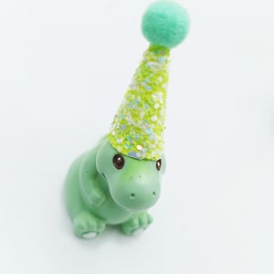 May include: A green dinosaur figurine wearing a green and white glittery party hat with a green pom-pom on top.