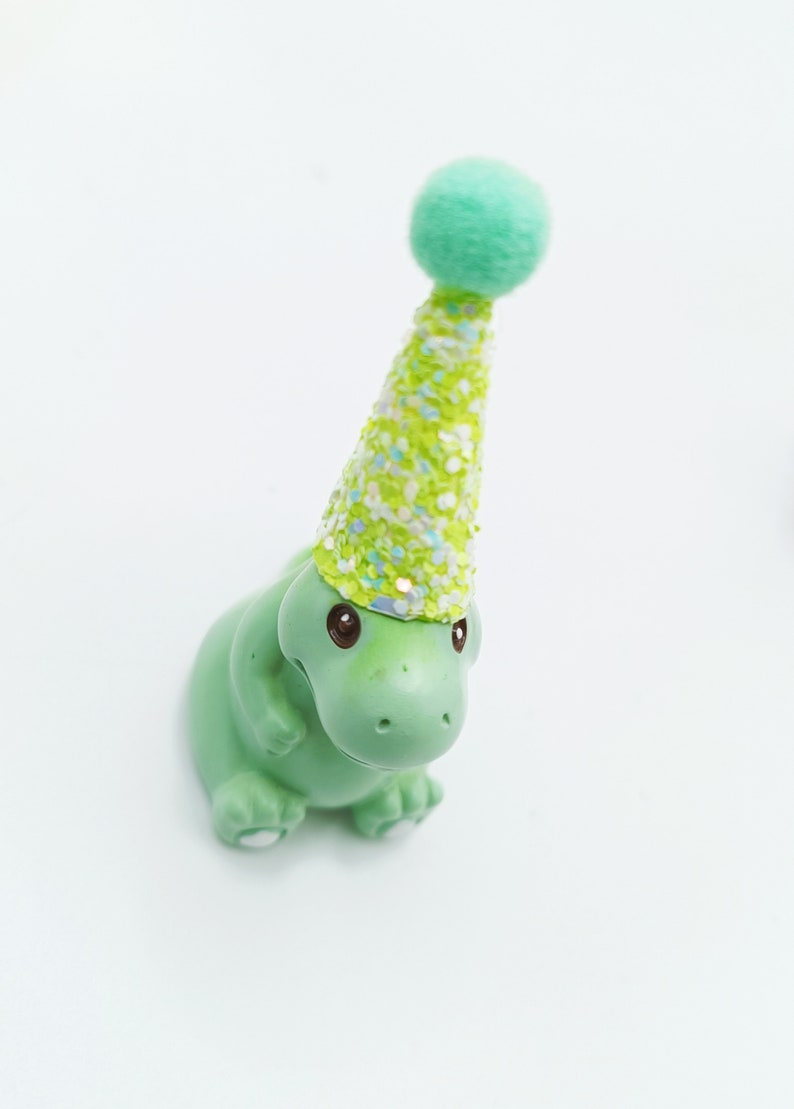 May include: A green dinosaur figurine wearing a green and white glittery party hat with a green pom-pom on top.