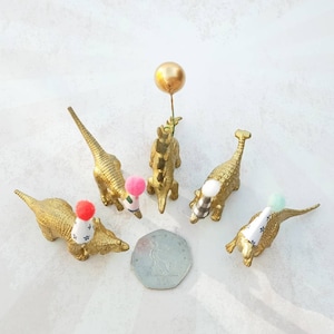 Puede incluir: Cinco figuritas de dinosaurio doradas que llevan sombreros de fiesta. Cada dinosaurio tiene un pomp&oacute;n de color diferente en la parte superior de su sombrero. Un dinosaurio sostiene un globo dorado.