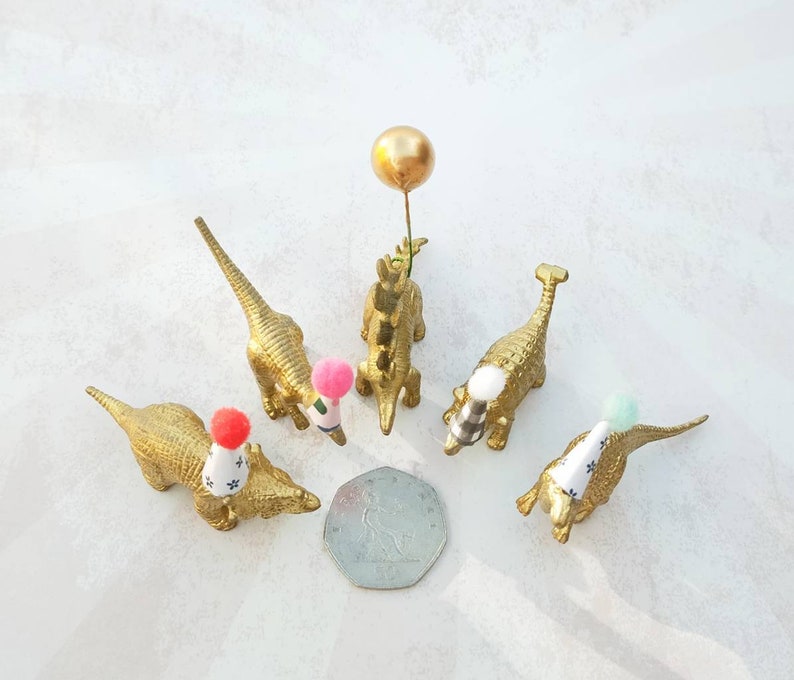 Puede incluir: Cinco figuritas de dinosaurio doradas que llevan sombreros de fiesta. Cada dinosaurio tiene un pomp&oacute;n de color diferente en la parte superior de su sombrero. Un dinosaurio sostiene un globo dorado.