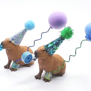 May include: Two brown capybara figurines wearing blue and green party hats with blue and green balloons attached to their hats.