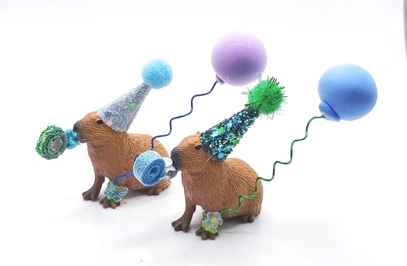 May include: Two brown capybara figurines wearing blue and green party hats with blue and green balloons attached to their hats.