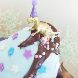 Puede incluir: Un pastel azul y blanco decorado con glaseado de chocolate, chispas y estrellas. Un peque&ntilde;o dinosaurio dorado con un gorro de fiesta morado se sienta en la parte superior del pastel.