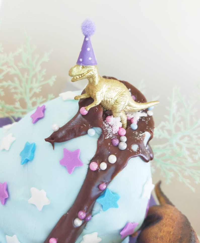 Puede incluir: Un pastel azul y blanco decorado con glaseado de chocolate, chispas y estrellas. Un peque&ntilde;o dinosaurio dorado con un gorro de fiesta morado se sienta en la parte superior del pastel.