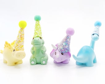 May include: Four colorful dinosaur figurines wearing party hats. The dinosaurs are green, purple, blue, and yellow. The hats are decorated with glitter.