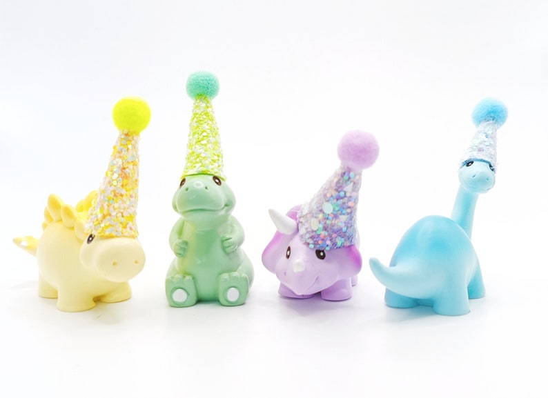 May include: Four colorful dinosaur figurines wearing party hats. The dinosaurs are green, purple, blue, and yellow. The hats are decorated with glitter.