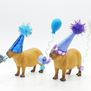 May include: Two capybaras wearing party hats and blowing party favours. One capybara is wearing a blue hat with a blue pom-pom and the other is wearing a purple hat with a purple pom-pom. Both capybaras are holding blue and purple party favours in their mouths.