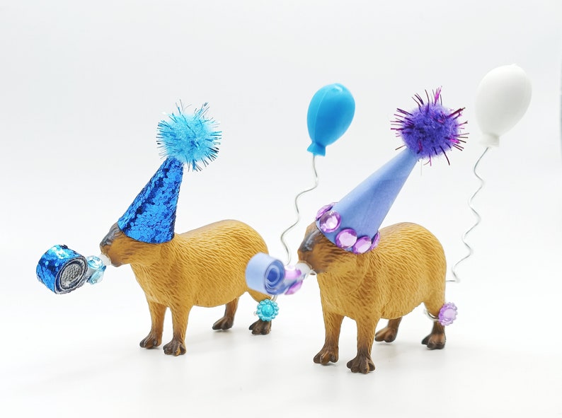 May include: Two capybaras wearing party hats and blowing party favours. One capybara is wearing a blue hat with a blue pom-pom and the other is wearing a purple hat with a purple pom-pom. Both capybaras are holding blue and purple party favours in their mouths.