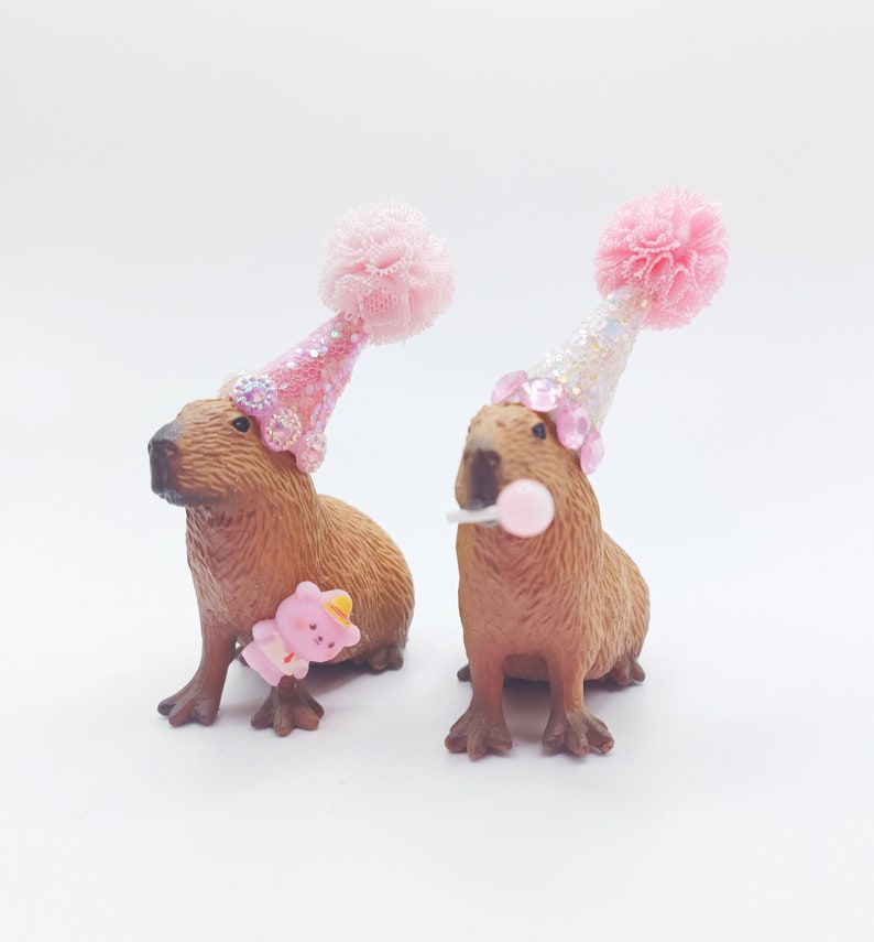 May include: Two brown capybara figurines wearing pink and white party hats with glitter and pom-poms. One capybara is holding a pink lollipop and the other is holding a pink teddy bear.